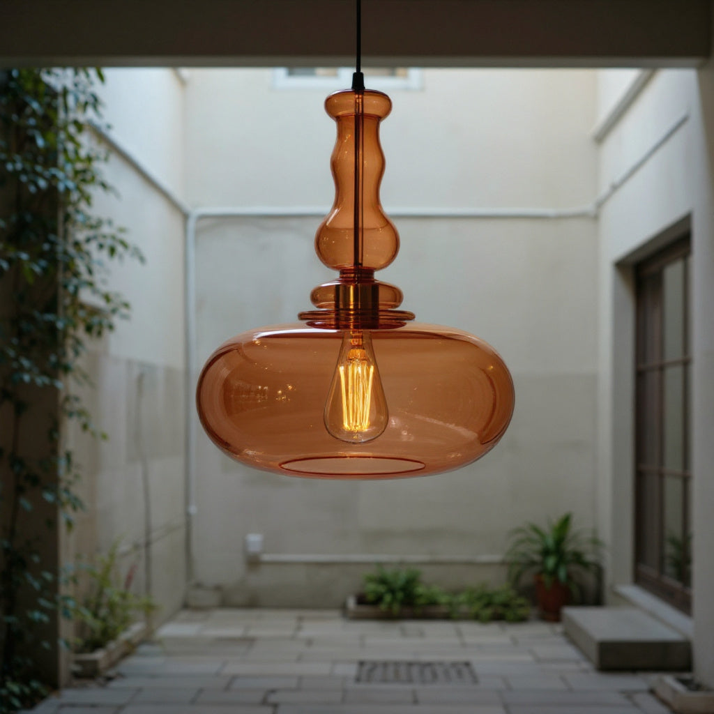 Amber glass pendant light hanging in an outdoor setting with plants and a wall.